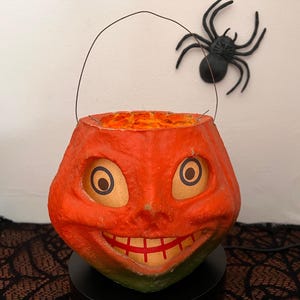 Puede incluir: Una calabaza de Halloween vintage de papel maché naranja con una sonrisa de dientes y ojos negros. La calabaza tiene un asa de alambre y está sentada sobre una base negra.