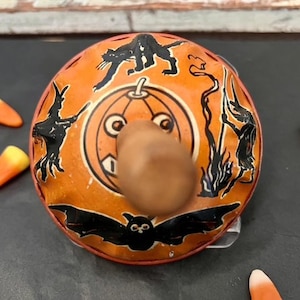 May include: A round orange metal spinning top toy featuring black illustrations of a cat, a bat, and a witch stirring a cauldron around a central jack-o'-lantern face. A brown wooden handle protrudes from the centre of the toy.