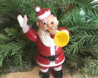 May include: A vintage plastic Santa Claus figurine playing a yellow trumpet. He is wearing a red and white suit with a white beard and black boots.