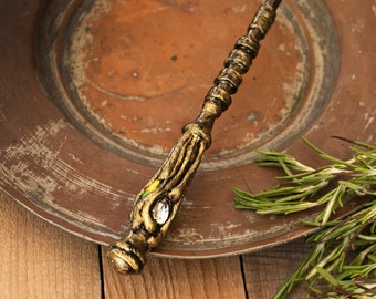 Op de afbeelding: Een gouden en bruine houten toverstok met een heldere edelsteen aan het uiteinde. De toverstok ligt op een koperen bord met een takje rozemarijn.