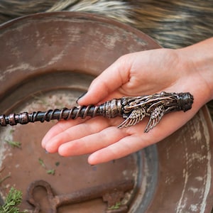 May include: A dark brown and copper-colored wand with a detailed winged design at the top. The wand is held in a hand, resting on a copper-colored plate.