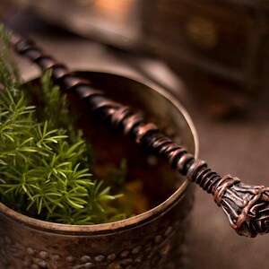 May include: A copper-colored metal wand with a rose-shaped tip, resting in a copper-colored metal cup with green foliage.