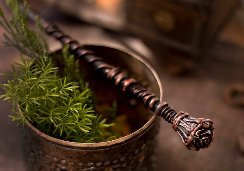 May include: A copper-colored metal wand with a rose-shaped tip, resting in a copper-colored metal cup with green foliage.
