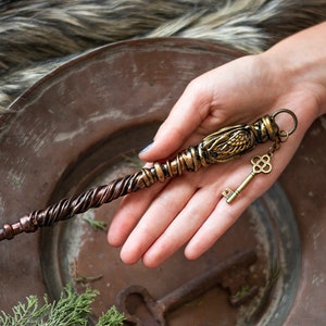 May include: A gold and copper wand with a key charm. The wand has a twisted copper base and a gold ornate top with a winged design. The key charm is attached to the wand with a gold chain.