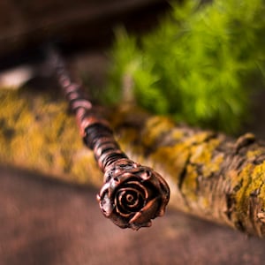 May include: A copper-colored wand with a rose-shaped design at the end. The wand is lying on a wooden surface with moss.