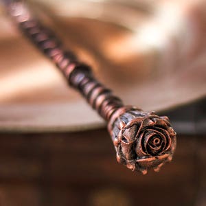 May include: A copper-colored wand with a detailed rose design at the tip. The wand is made of wire wrapped around a wooden core.