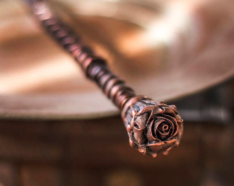 May include: A copper-colored wand with a detailed rose design at the tip. The wand is made of wire wrapped around a wooden core.