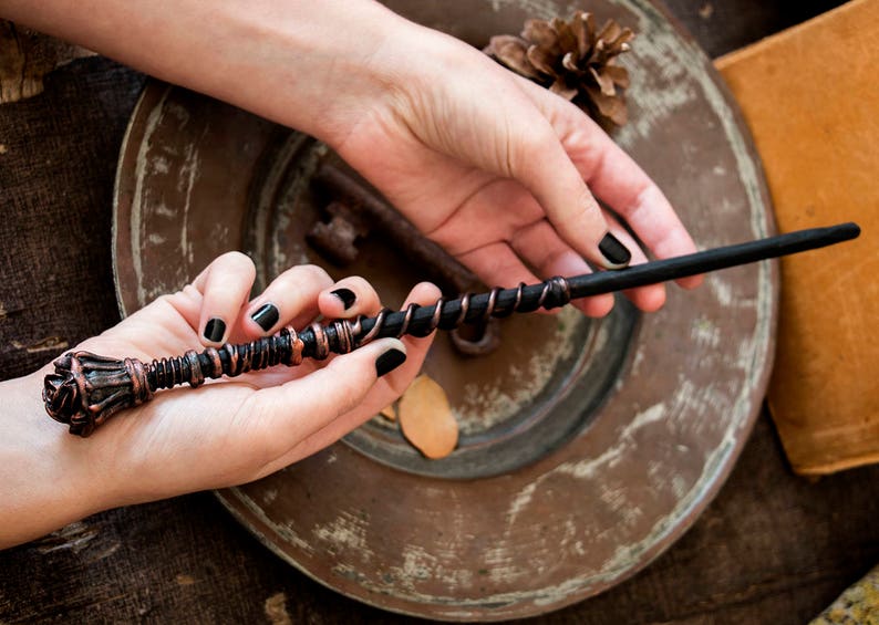 May include: A hand holds a black and copper wand with a rose-shaped handle. The wand is on a wooden surface with a key and a pine cone.