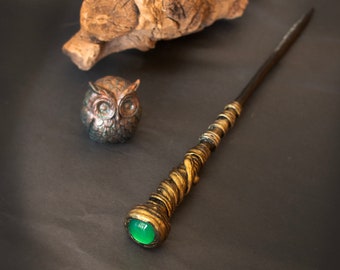 Op de afbeelding: Een decoratieve toverstaf met een groene edelsteenpunt en gouden en zwarte accenten. De toverstaf ligt op een donkergrijs oppervlak naast een klein uilbeeldje.