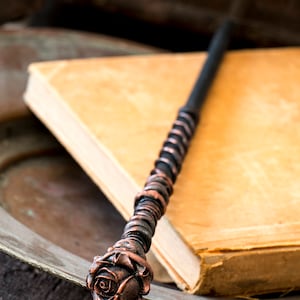 May include: A black and copper wand with a rose-shaped handle. The wand is lying on top of a closed book.