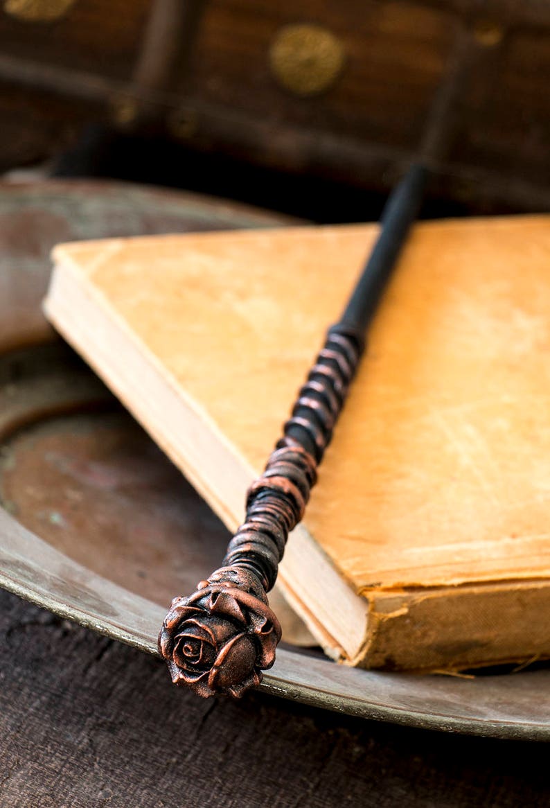 May include: A black and copper wand with a rose-shaped handle. The wand is lying on top of a closed book.