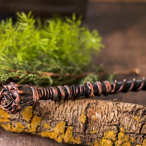 May include: A dark brown and copper colored wand with a rose-shaped handle. The wand is lying on a piece of bark with green foliage in the background.