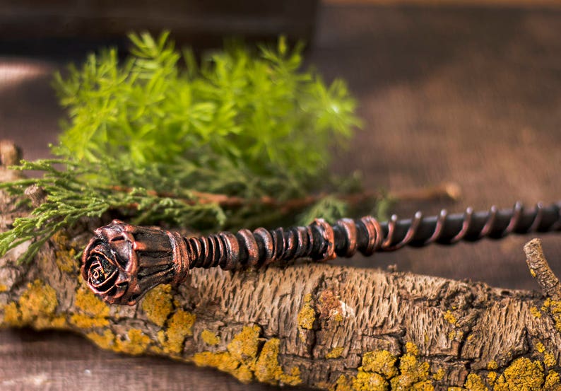 May include: A dark brown and copper colored wand with a rose-shaped handle. The wand is lying on a piece of bark with green foliage in the background.
