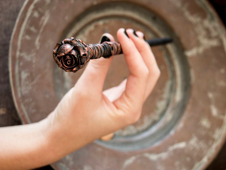 May include: A copper-colored rose-shaped wand with a black handle. The wand is held in a hand.