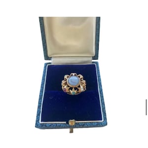May include: A gold ring with a large, oval, light blue center stone, surrounded by smaller gemstones and star-shaped accents. The ring is displayed in a blue velvet-lined jewelry box with a gold clasp.