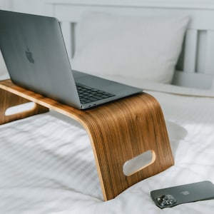 Peut inclure: Un ordinateur portable sur un support pour ordinateur portable en bois, avec un smartphone sur un couvre-lit blanc.