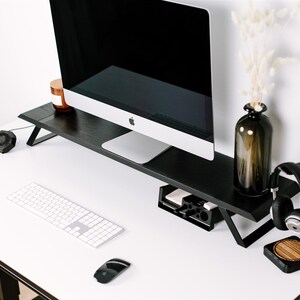 May include: A black computer monitor stand with a white keyboard, a black mouse, and a black headset stand with a wooden base on a white desk.