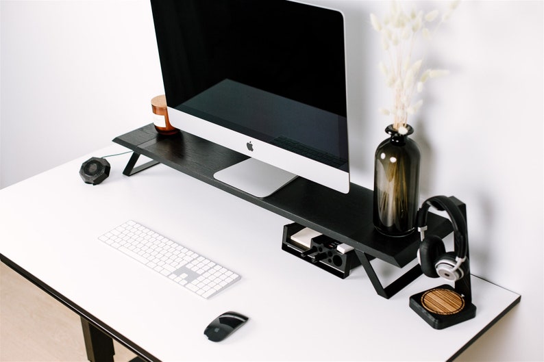 May include: A black computer monitor stand with a white keyboard, a black mouse, and a black headset stand with a wooden base on a white desk.