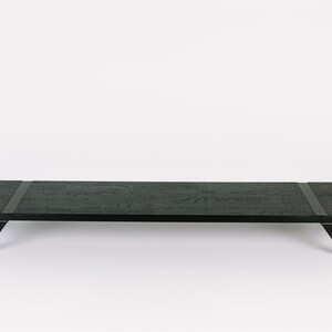 May include: A long, black wooden coffee table with a rectangular top and angled legs. The table has a dark wood grain and two black leather straps across the top.