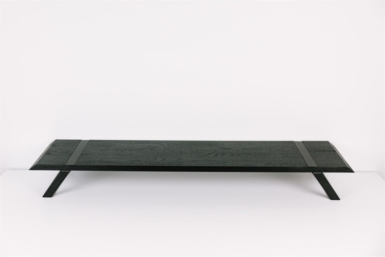 May include: A long, black wooden coffee table with a rectangular top and angled legs. The table has a dark wood grain and two black leather straps across the top.