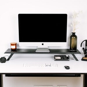 May include: A white desk with a black monitor stand, a white keyboard, a black mouse, a black speaker, a black headset stand with a black headset, a brown candle, and a brown vase with dried white flowers.