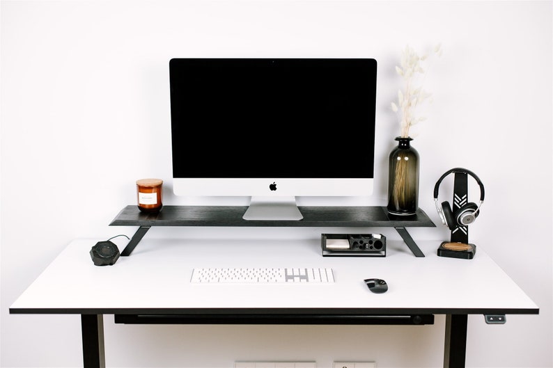 May include: A white desk with a black monitor stand, a white keyboard, a black mouse, a black speaker, a black headset stand with a black headset, a brown candle, and a brown vase with dried white flowers.