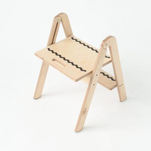 Può includere: Uno sgabello in legno color chiaro dal design semplice. Lo sgabello ha due gradini, ciascuno con un motivo a linea ondulata nera. Le gambe sono angolate per la stabilità. Una maniglia ritagliata è sul gradino superiore.