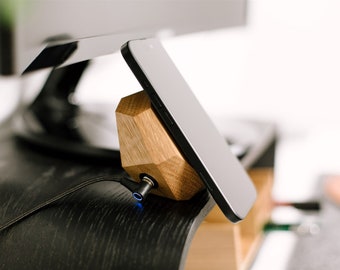 May include: A wooden phone stand with a geometric design holds a black smartphone. A black charging cable is plugged into the stand.