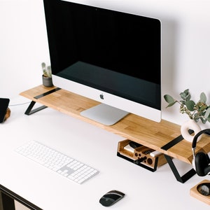 May include: A white desk with a wooden monitor stand, a white keyboard, a black mouse, a black phone, a black headphone stand with black headphones, a wooden organizer with pens and pencils, and a small potted plant.