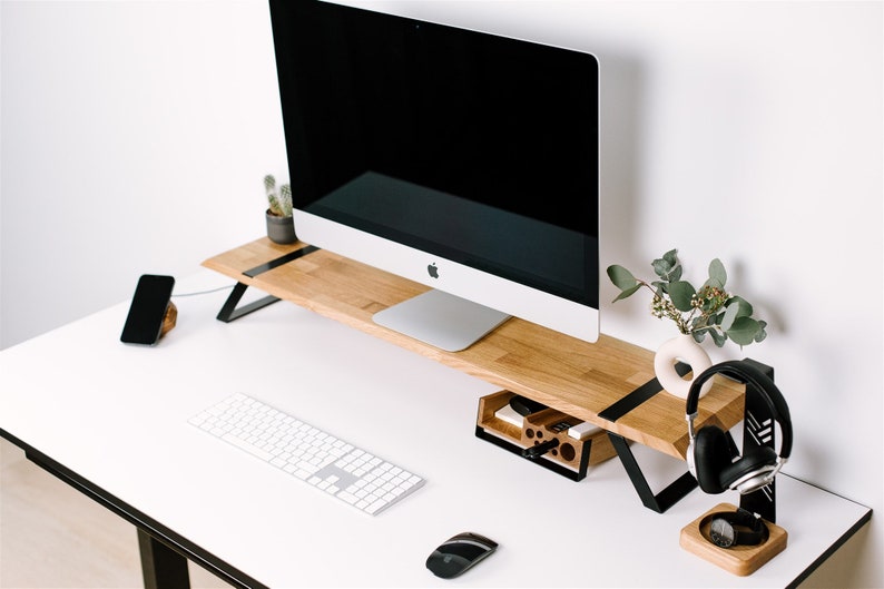 May include: A white desk with a wooden monitor stand, a white keyboard, a black mouse, a black phone, a black headphone stand with black headphones, a wooden organizer with pens and pencils, and a small potted plant.
