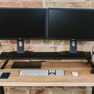 May include: A wooden desk with a black metal frame. Two black computer monitors are on a black monitor stand. A white keyboard, a white mouse, a black cell phone, and a pair of white headphones are on the desk.