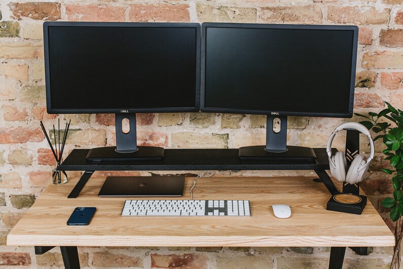May include: A wooden desk with a black metal frame. Two black computer monitors are on a black monitor stand. A white keyboard, a white mouse, a black cell phone, and a pair of white headphones are on the desk.