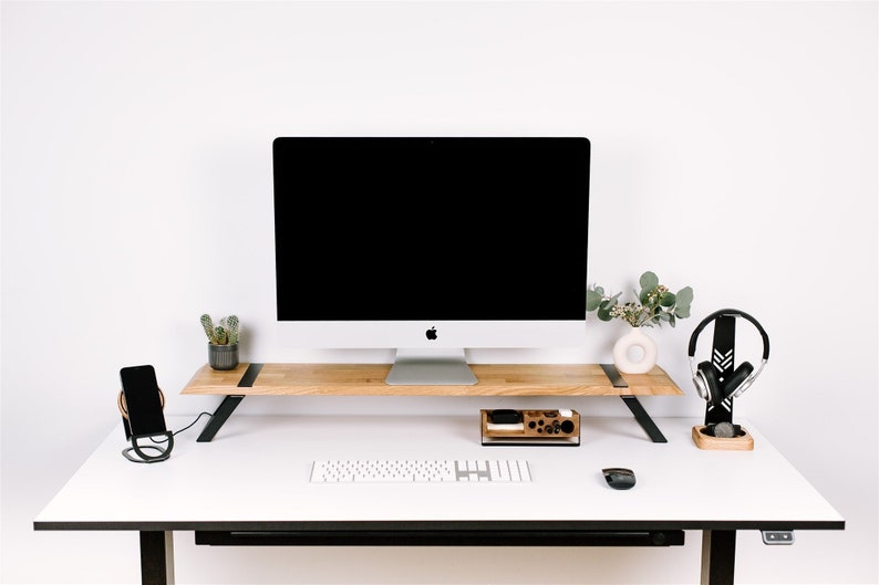 May include: A white desk with a black monitor stand, a black keyboard, a black mouse, a black phone stand, a black headphone stand, a wooden organizer, a small potted cactus, and a vase with green leaves.