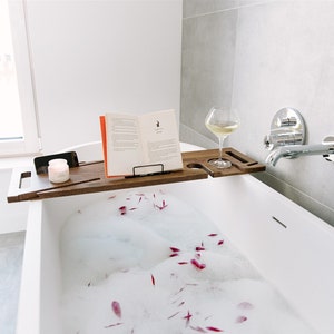 Puede incluir: Una bandeja de baño de madera con un libro, una copa de vino y una vela. La bandeja está colocada en el borde de una bañera blanca llena de burbujas y pétalos de flores rosas.