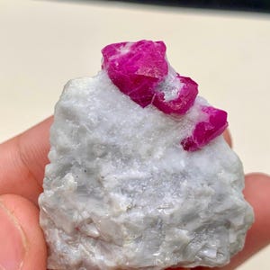 May include: A cluster of bright pink ruby crystals embedded in a white matrix. The ruby crystals are sharp and well-defined, with a glassy luster. The matrix is rough and textured.