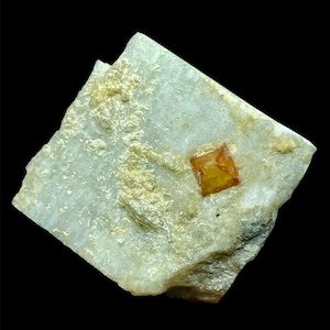 Puede incluir: Un pequeño cristal cúbico de color marrón anaranjado incrustado en una matriz blanca, gris y dorada. El cristal es translúcido y tiene un ligero brillo.