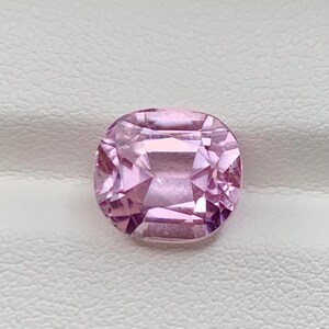 May include: A faceted, cushion-cut gemstone in a soft pink hue. The gem has a symmetrical design with multiple facets that reflect light. The stone is set against a white, textured background.