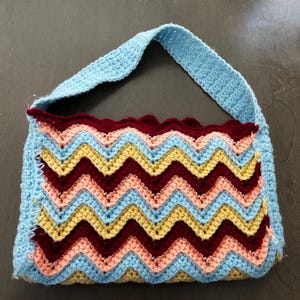 Puede incluir: Bolso de crochet hecho a mano con correa y cuerpo azul claro. El bolso presenta un colorido patrón de chevrón en tonos azul claro, melocotón, amarillo y burdeos. El borde superior está acabado con un ribete burdeos festoneado.