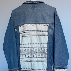 Könnte beinhalten: Blaue Jeansjacke mit einem weißen Einsatz auf dem Rücken, verziert mit schwarzer geometrischer Stickerei. Die Jacke hat einen klassischen Kragen, geknöpfte Manschetten und einen Used-Look. Sie hängt an einem weißen Kleiderbügel.