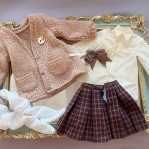 May include: A brown knitted cardigan with a cat pin, a white button-down shirt with a brown bow, a brown plaid skirt, and a white crocheted scarf.