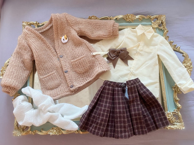 May include: A brown knitted cardigan with a cat pin, a white button-down shirt with a brown bow, a brown plaid skirt, and a white crocheted scarf.