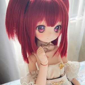 Op de afbeelding: Een pop met lang, rood haar dat in paardenstaarten is gestyled. De pop draagt een witte jurk met een gouden riem en een parelketting. De pop heeft roze oogschaduw en blauwe ogen. De pop glimlacht en kijkt naar rechts. De tekst "Strawberry Ruby" staat onderaan de afbeelding.