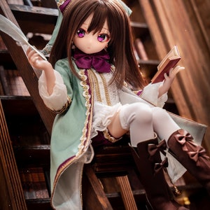 Puede incluir: Una muñeca con un vestido verde y dorado con un lazo morado, sentada en una escalera de madera frente a una estantería. La muñeca sostiene un libro y una pluma.