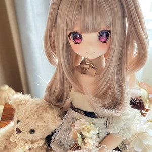 Puede incluir: Una muñeca con el pelo largo y castaño claro recogido en dos coletas con lazos blancos. La muñeca tiene ojos grandes, rosa y morado, y lleva un vestido blanco y marrón con una capa de encaje. La muñeca lleva un pequeño osito de peluche marrón.