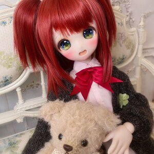 May include: A red-haired doll with green eyes is sitting on a white chair holding a beige teddy bear. The doll is wearing a black cardigan with a green clover pin, a pink shirt, and a red bow.