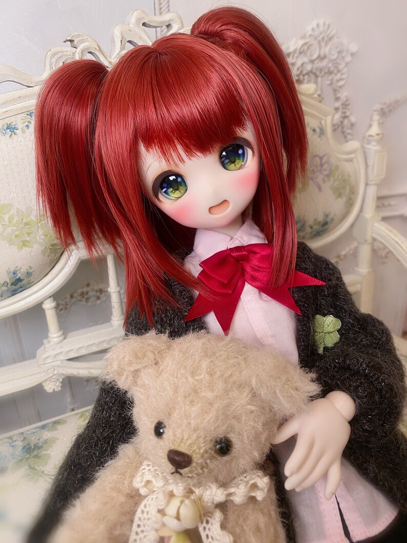 May include: A red-haired doll with green eyes is sitting on a white chair holding a beige teddy bear. The doll is wearing a black cardigan with a green clover pin, a pink shirt, and a red bow.