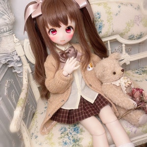 May include: A doll with brown hair in pigtails wearing a brown cardigan, a white shirt, a plaid skirt, and white socks. The doll is sitting on a floral patterned chair with a teddy bear beside it.