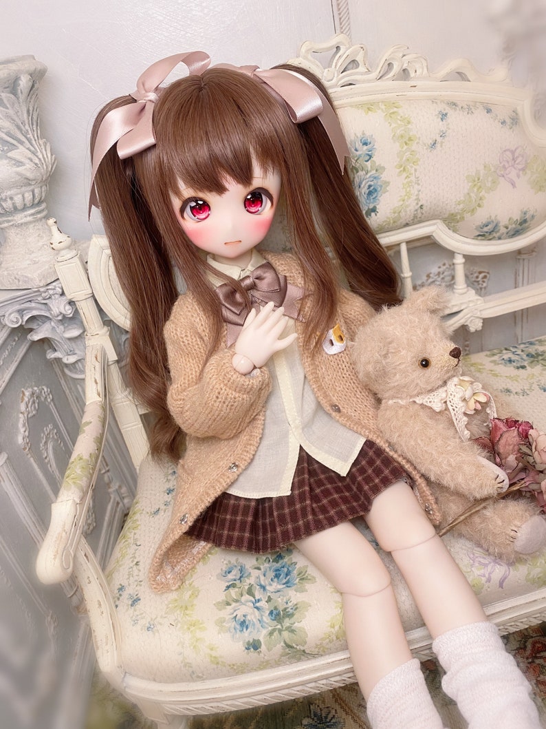 May include: A doll with brown hair in pigtails wearing a brown cardigan, a white shirt, a plaid skirt, and white socks. The doll is sitting on a floral patterned chair with a teddy bear beside it.