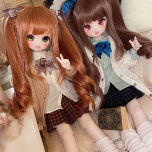 May include: Two dolls with long brown and orange hair wearing white shirts, brown sweaters, and plaid skirts. The dolls are sitting on a white couch with a book in front of them.
