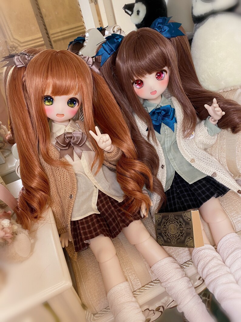 May include: Two dolls with long brown and orange hair wearing white shirts, brown sweaters, and plaid skirts. The dolls are sitting on a white couch with a book in front of them.